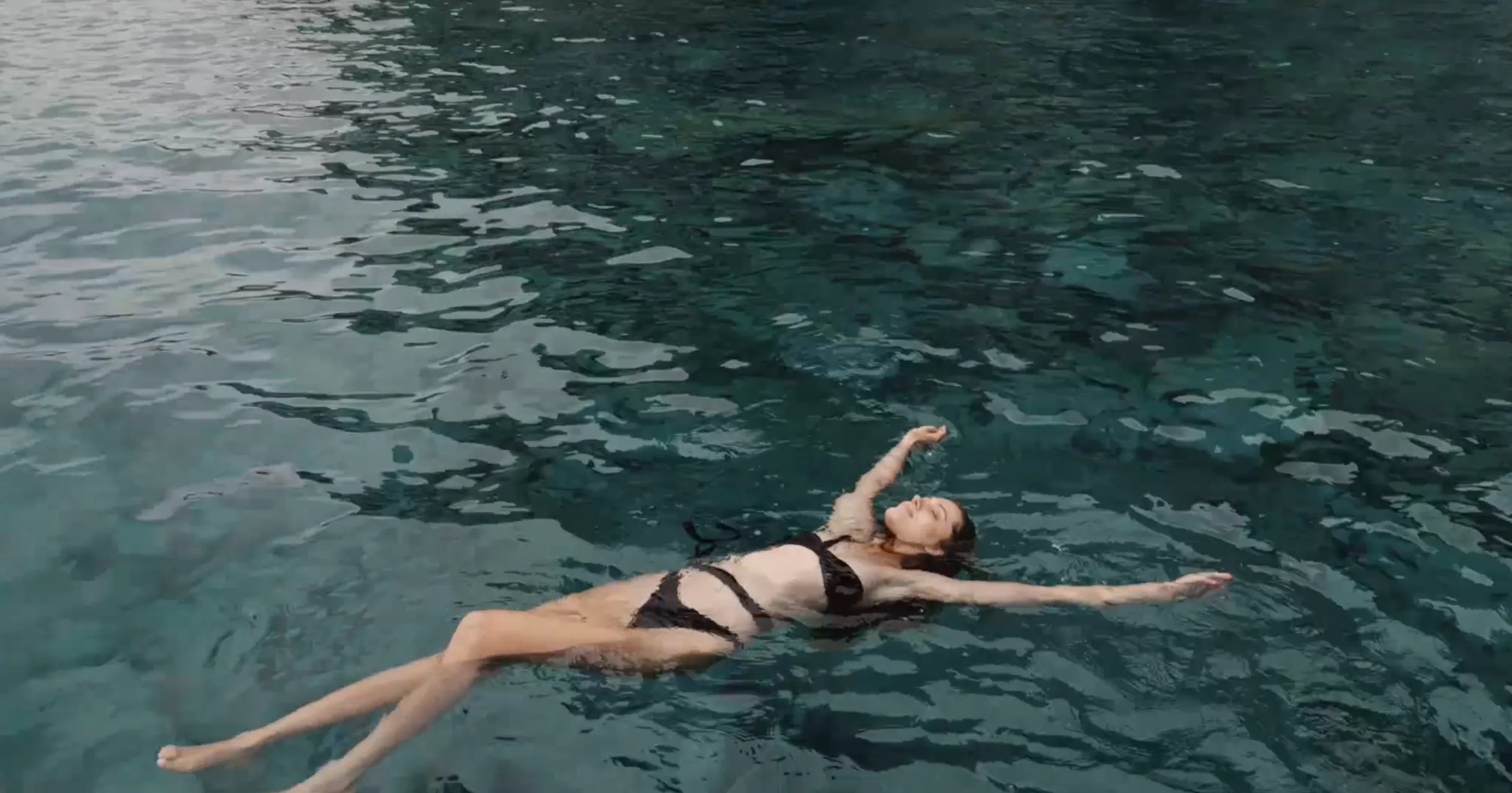 A woman in a black bikini floating on her back in clear, calm water with her arms and legs relaxed.