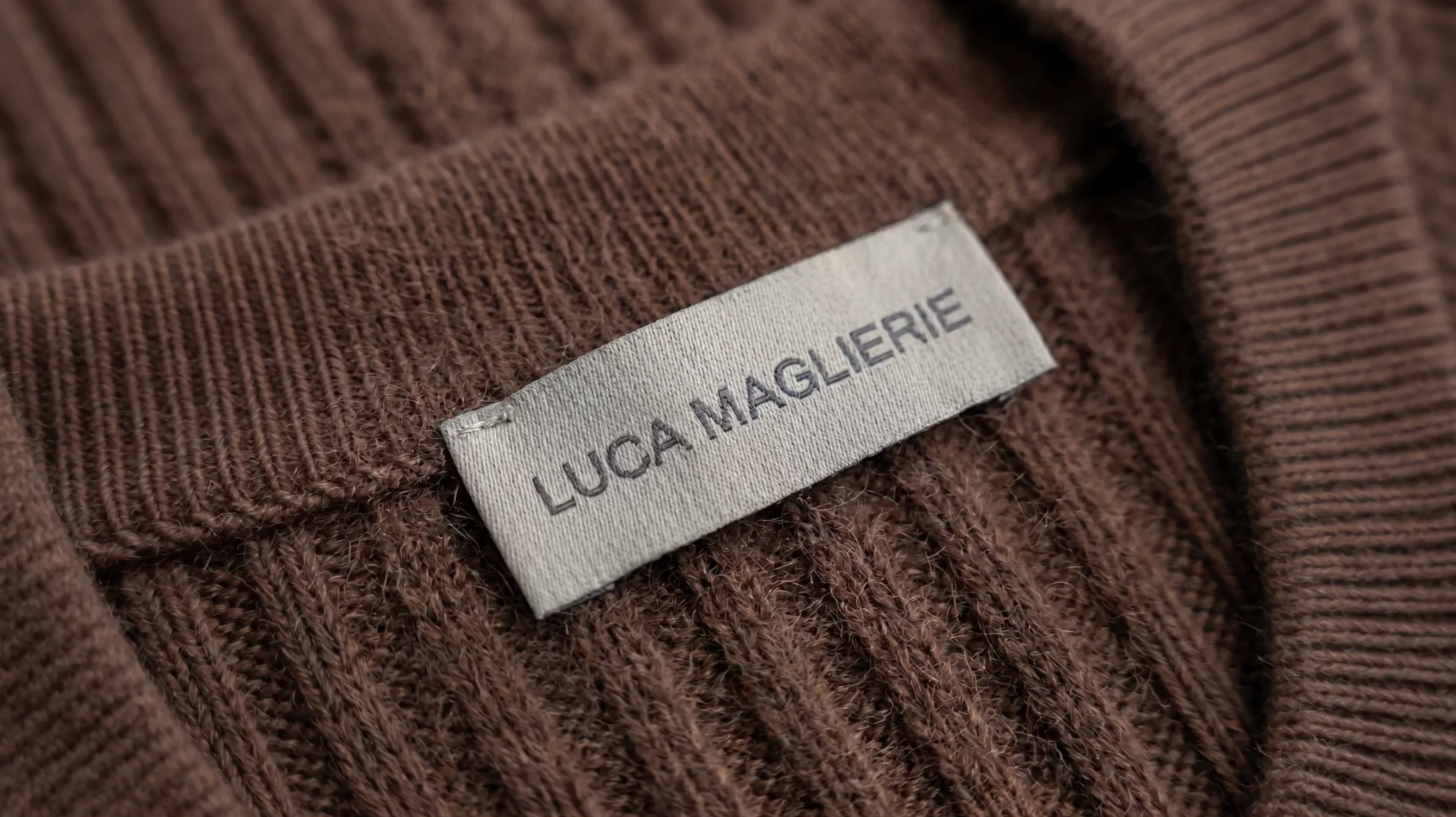 Close-up of a white clothing label sewn onto a brown knitted garment with ribbed texture, displaying the text 'LUCA MAGLIERIE' in uppercase letters.