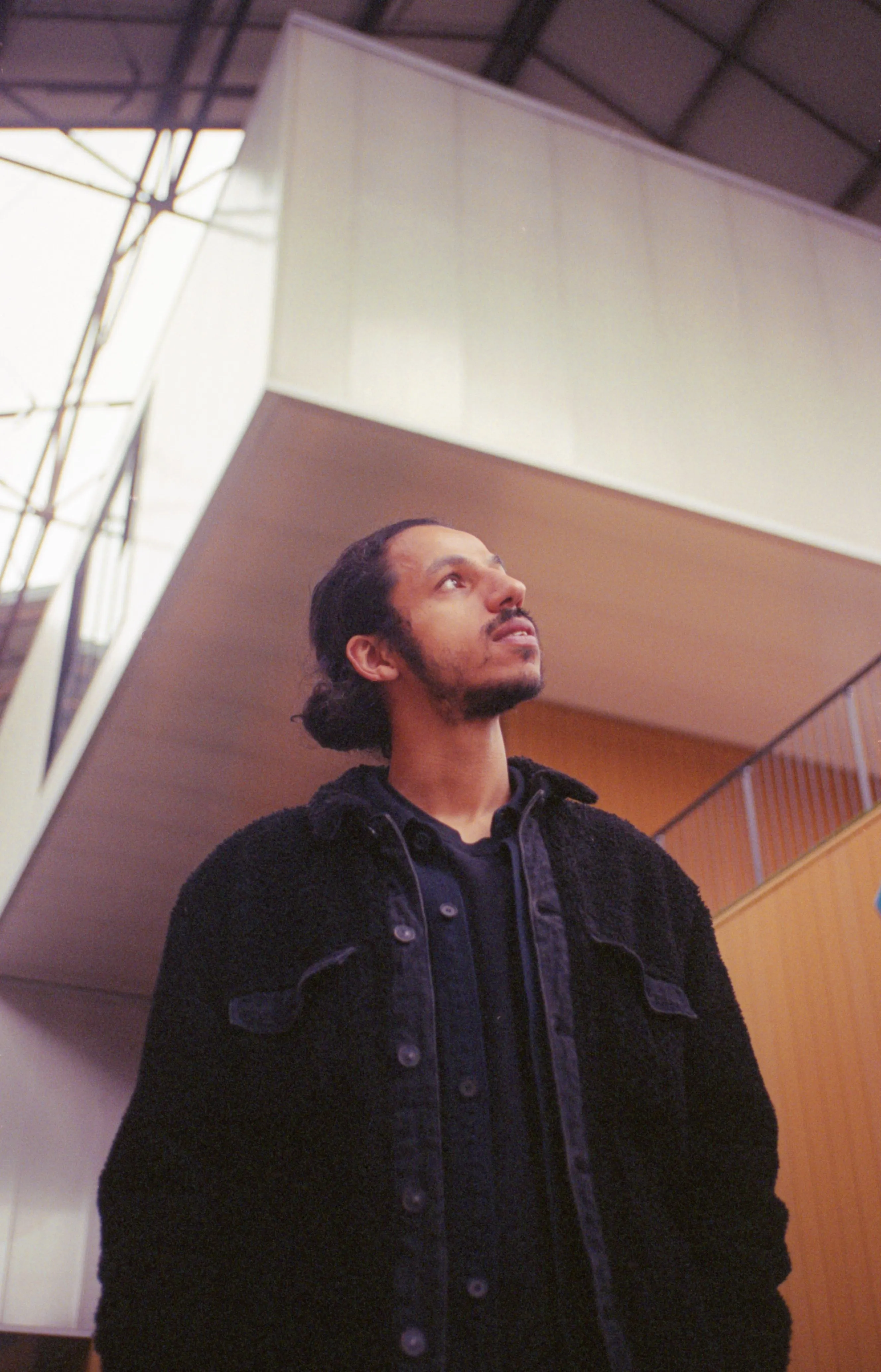 Man with a beard and tied-back hair wearing a black buttoned jacket, looking up indoors near a railing and modern ceiling.